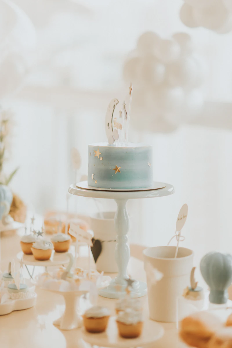 A pale blue birthday cake on a cream stand, with cupcakes behind it