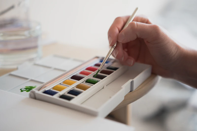 A hand dipping a brush into a box of watercolors